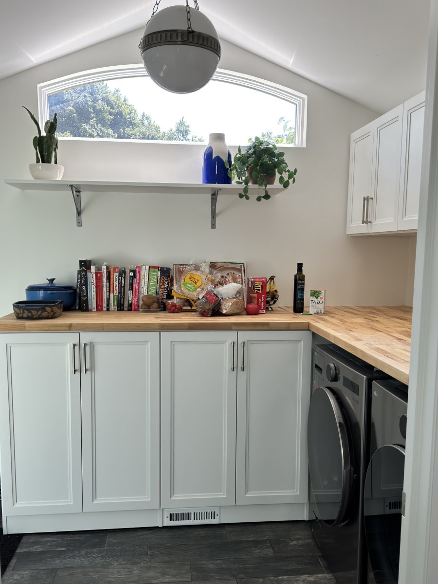 Small kitchen with white cabinets, books, plants, and groceries on a wooden countertop.