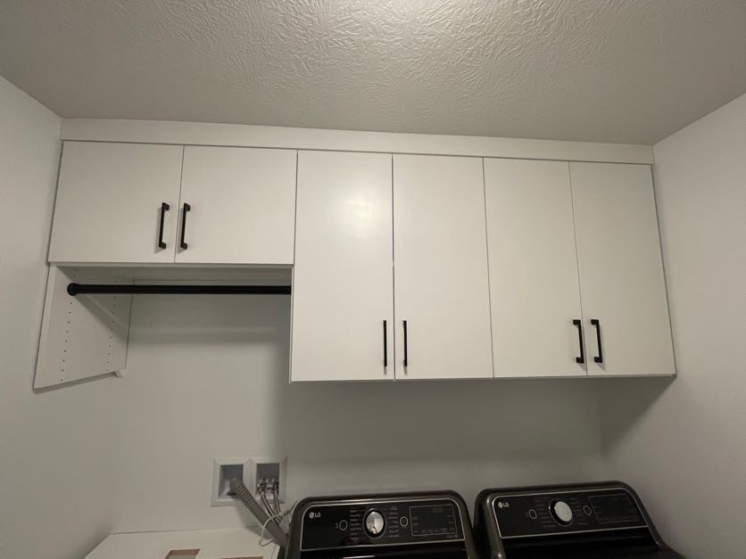 White cabinets with black handles are above two dark-colored washing machines in a laundry room.