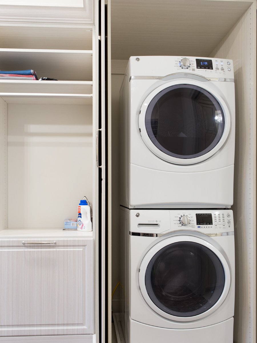 Stacked washer and dryer next to shelves and a cabinet in a laundry closet.