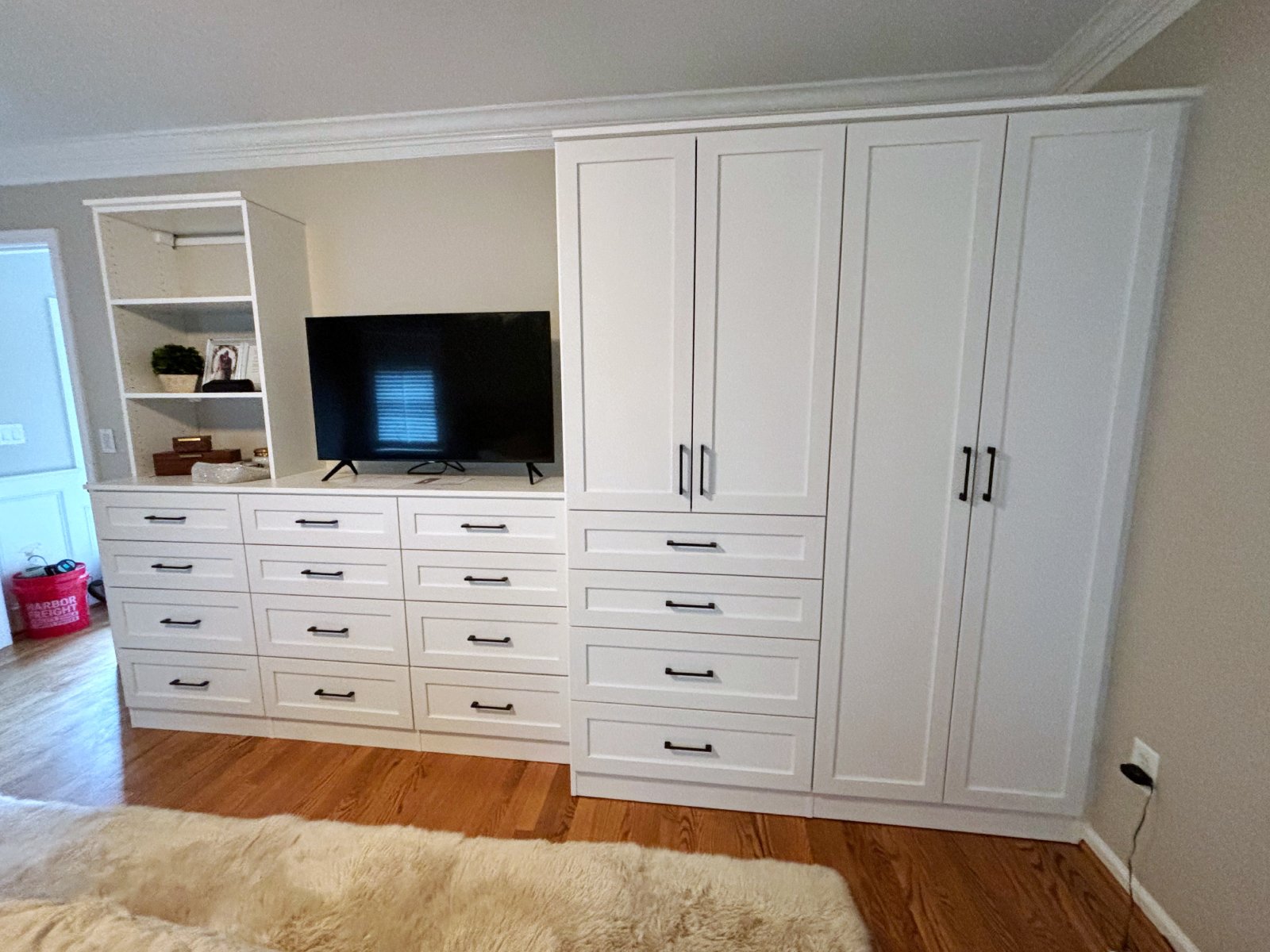 White built-in wardrobe with drawers, cabinets, shelves, and a TV, on a wooden floor.