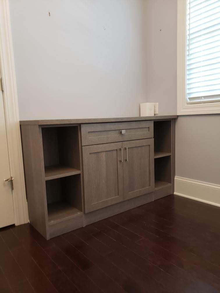 Gray wooden cabinet with open shelves stands against a light wall near a window and closed door.