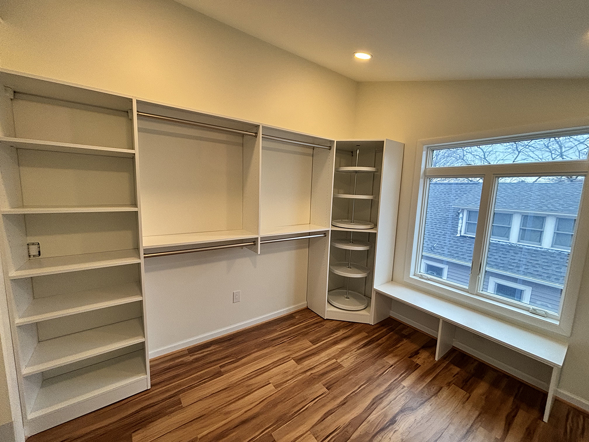 A walk-in closet with white shelves, hanging rods, corner shelves, and a window bench.