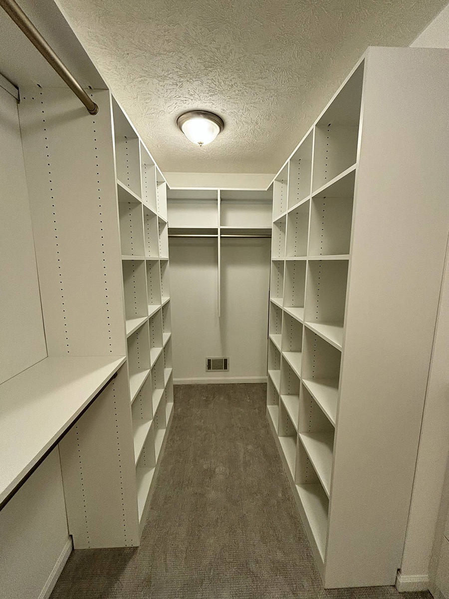 A walk-in closet with white shelves, hanging rods, and gray carpet under a ceiling light.