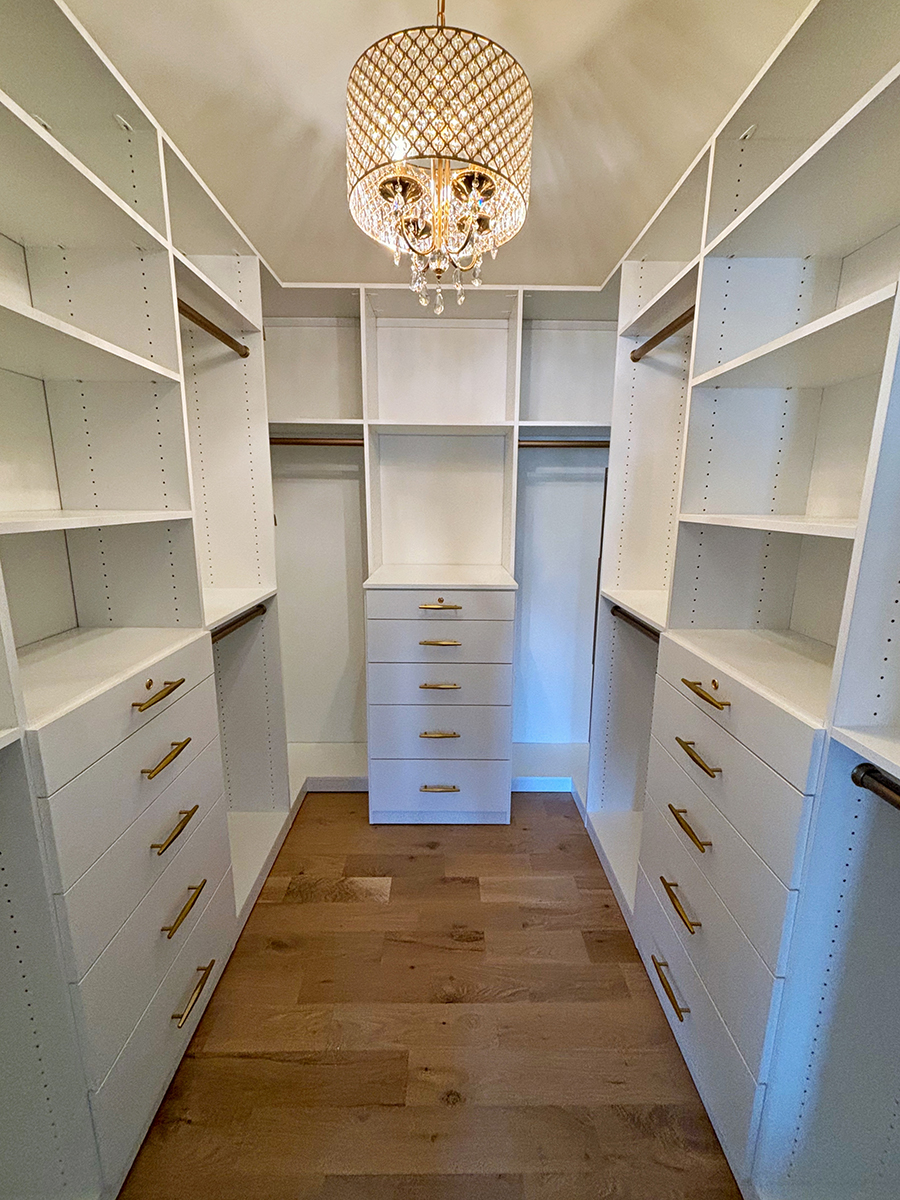 White walk-in closet with shelves, drawers, and gold handles, under a decorative chandelier.