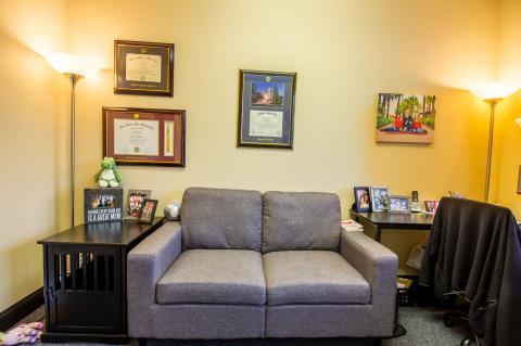 A small office with a gray loveseat, certificates on the wall, and photos on side tables.