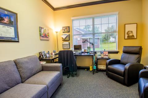 A cozy office with a sofa, recliner, desk, chair, and window overlooking a green lawn.