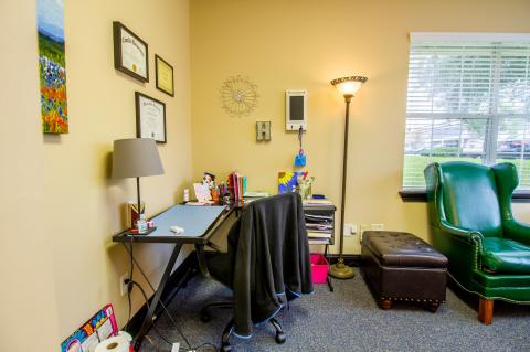 A small office with a desk, chair, lamp, green armchair, and window with blinds.