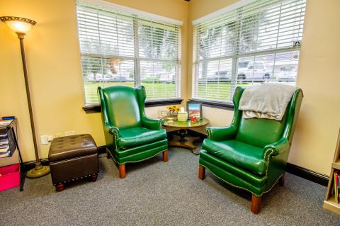 Two green armchairs sit by corner windows with a lamp, table, and ottoman nearby.