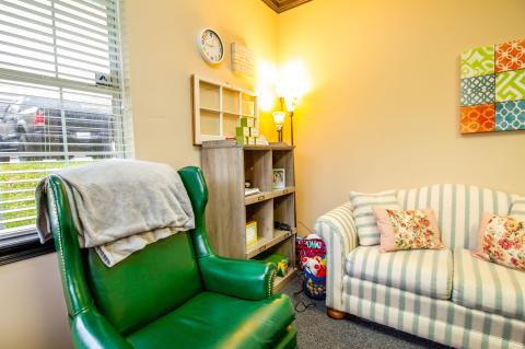 Cozy room with a green armchair, striped couch, bookshelf, lamp, and window with blinds.