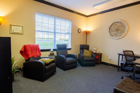 Cozy office with three recliners, a desk, large window, and warm lighting.