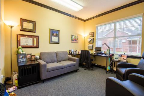 A cozy office with a gray sofa, desk, chair, certificates on the wall, and a large window.