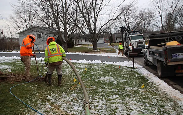 Workers in safety gear operate hoses on a snowy lawn near trucks and houses on a cold day.