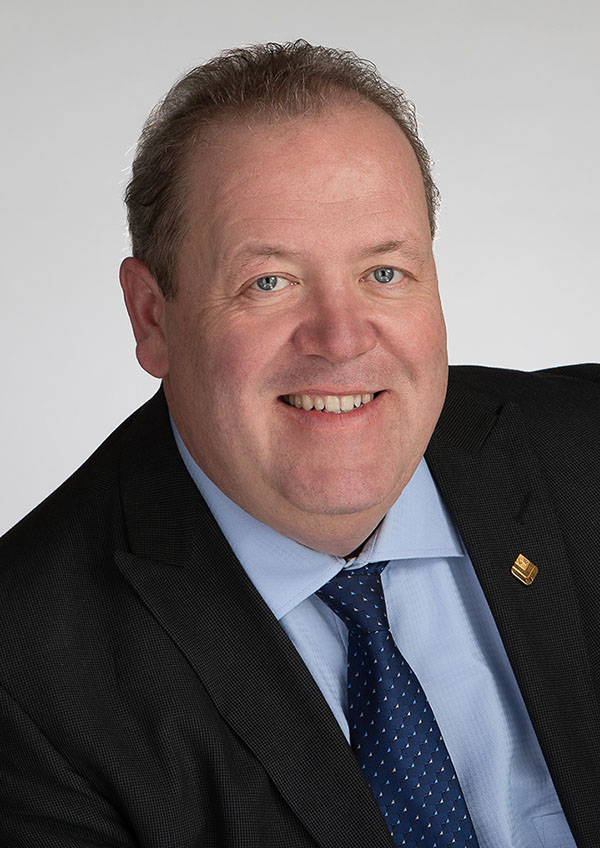 A man in a suit and blue tie smiles at the camera against a plain background.