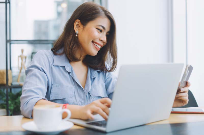 Smiling woman using a laptop at a cafe, holding a smartphone, with a cup of coffee on the table.