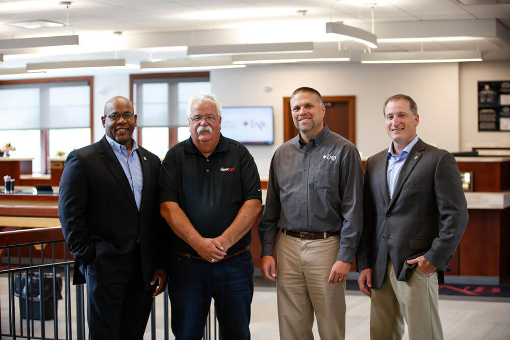 Four men standing in front of a desk in an office, discussing work projects and brainstorming ideas.