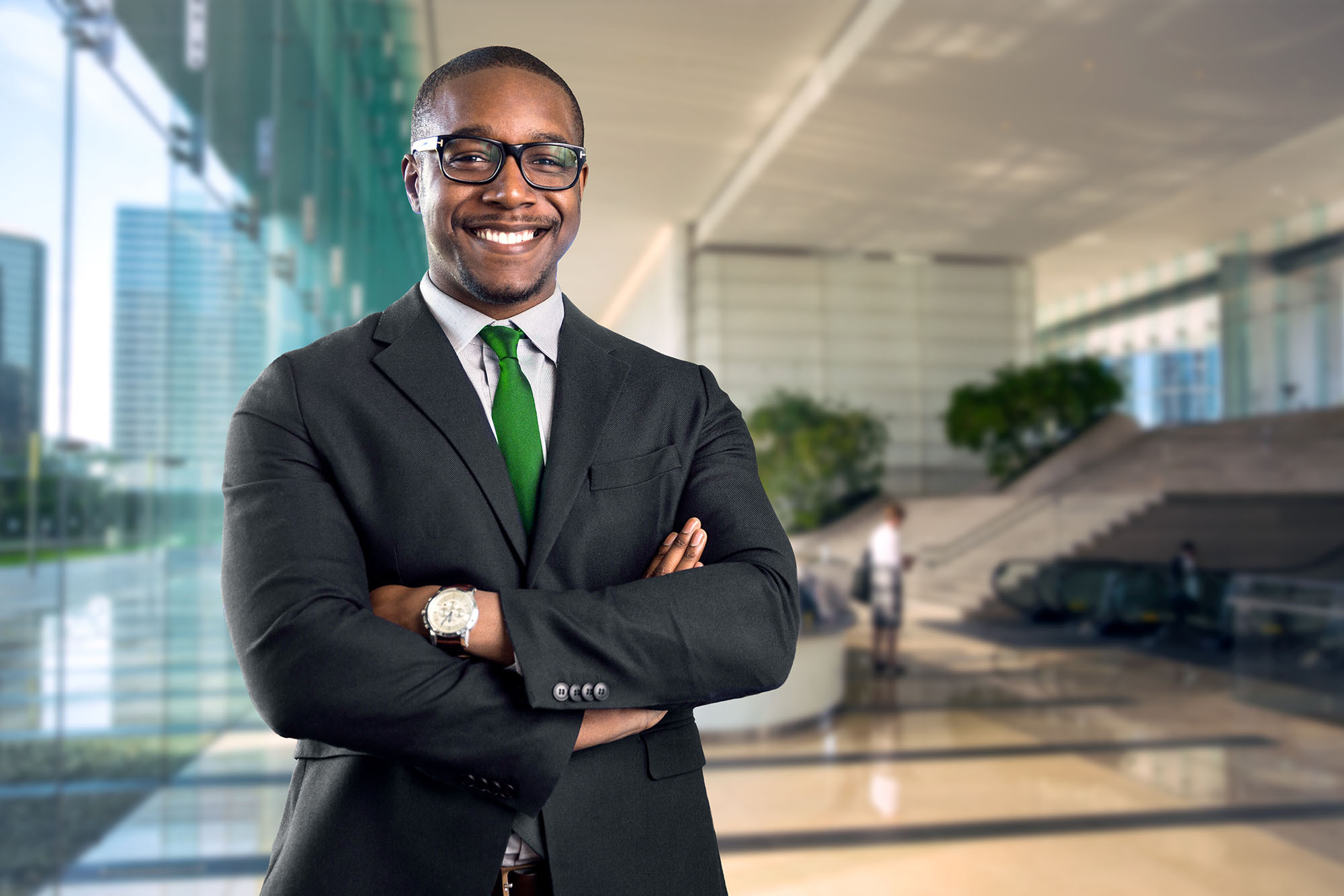 Smiling man in a suit with arms crossed stands in a bright, modern office building.
