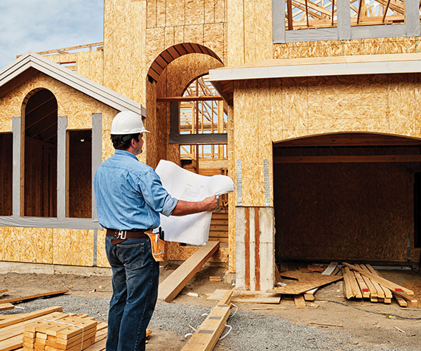 Man in hard hat examining blueprints at a wooden house construction site.