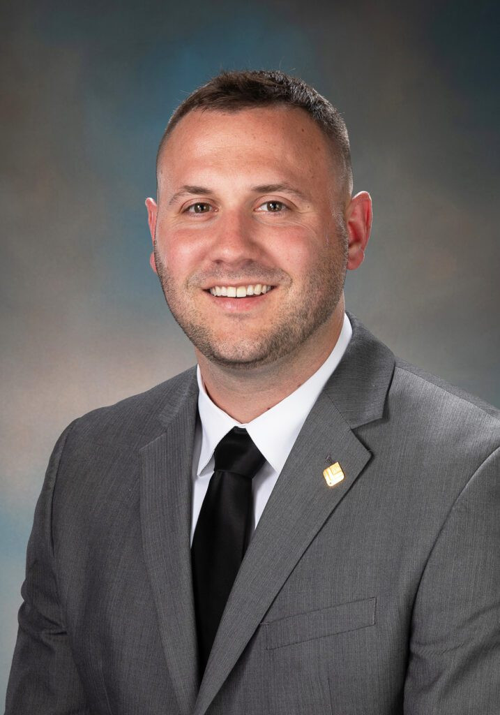 Smiling man in a gray suit and black tie posing for a professional portrait against a gray background.