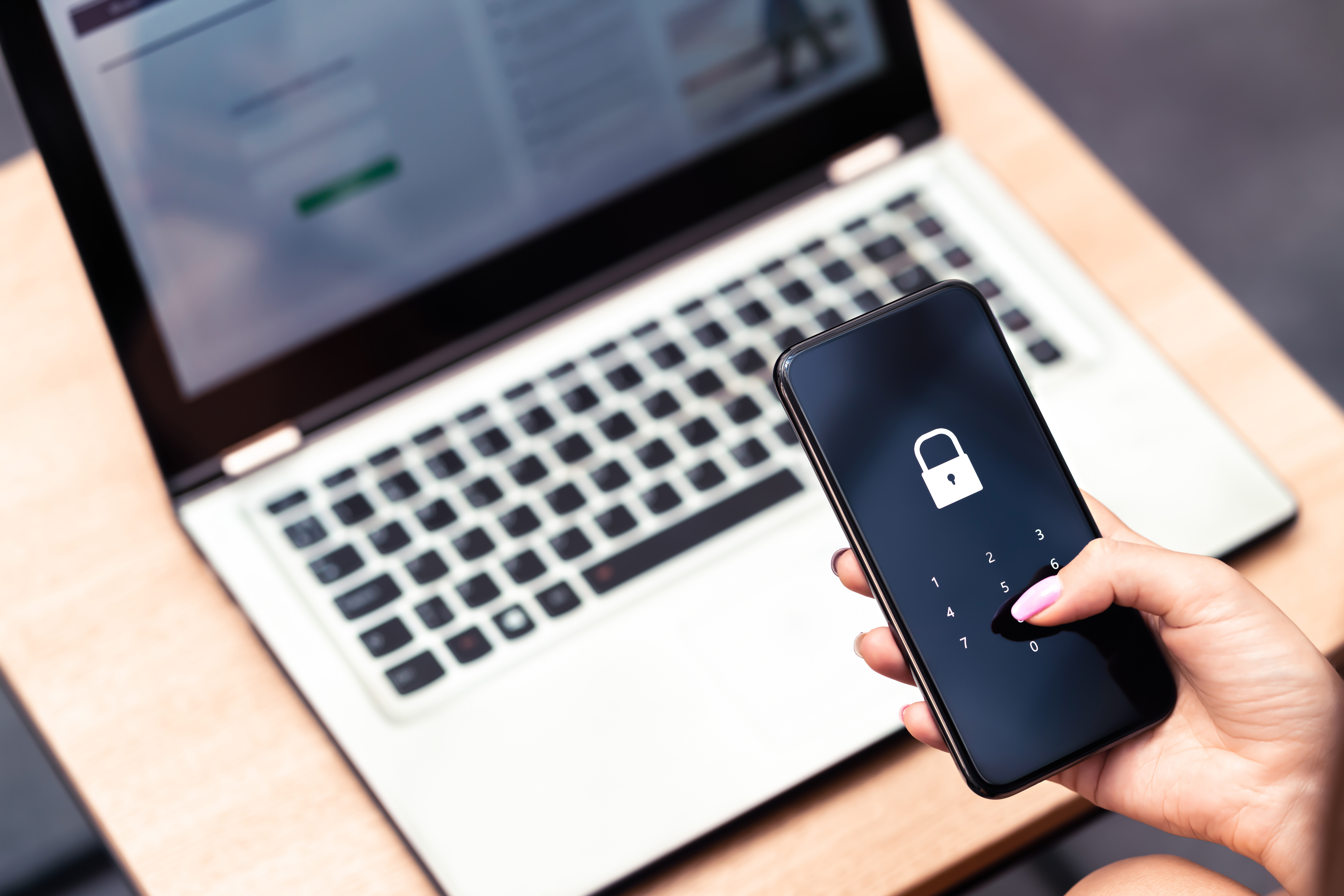 A person enters a passcode on a smartphone in front of a laptop displaying a login screen.