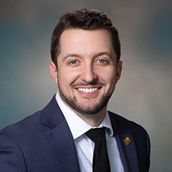 Smiling man in a dark suit, white shirt, and black tie against a blurred background.