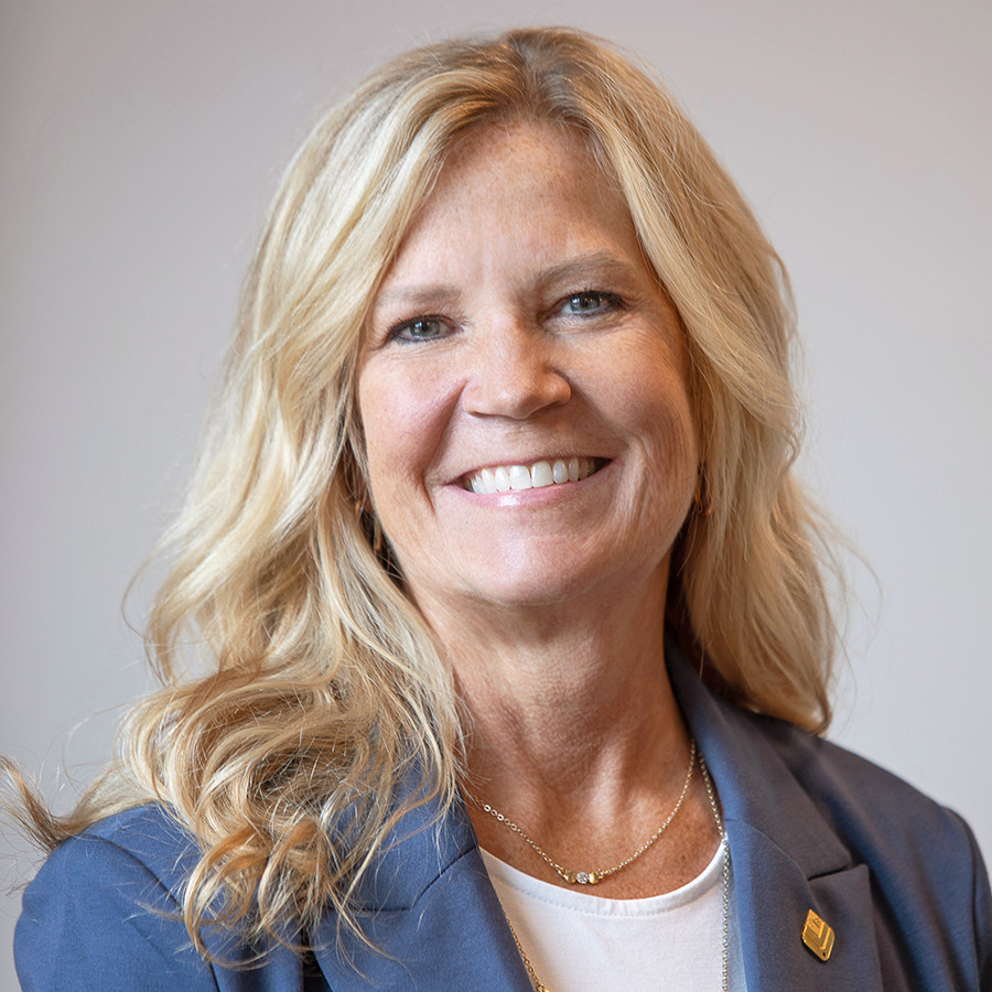 Smiling woman with long blonde hair wearing a blue blazer and white top against a neutral background.