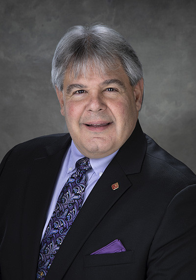 Smiling older man in a suit with a purple tie and pocket square, gray background.