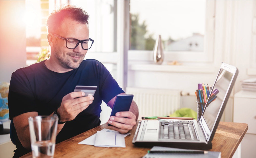 A man with glasses sitting at a desk, using a laptop and holding a credit card.