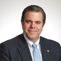 Man in a suit and blue tie smiling at the camera against a plain, light background.