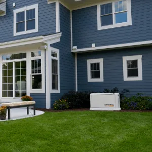 Blue house with white trim, green lawn, and a Generac generator beside a flowerbed.