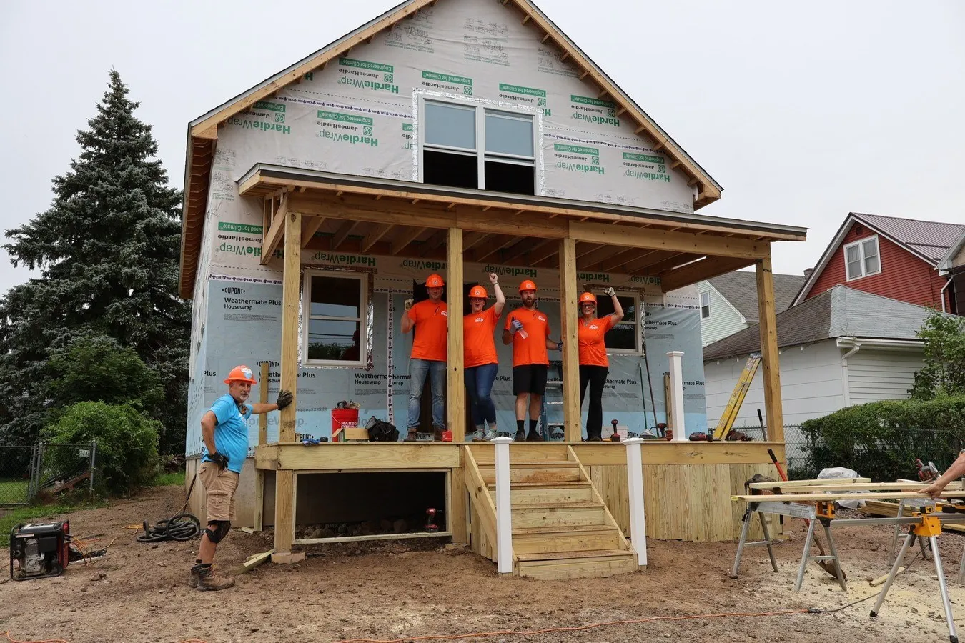 Five people in orange shirts and hard hats stand on the porch of a house under construction.