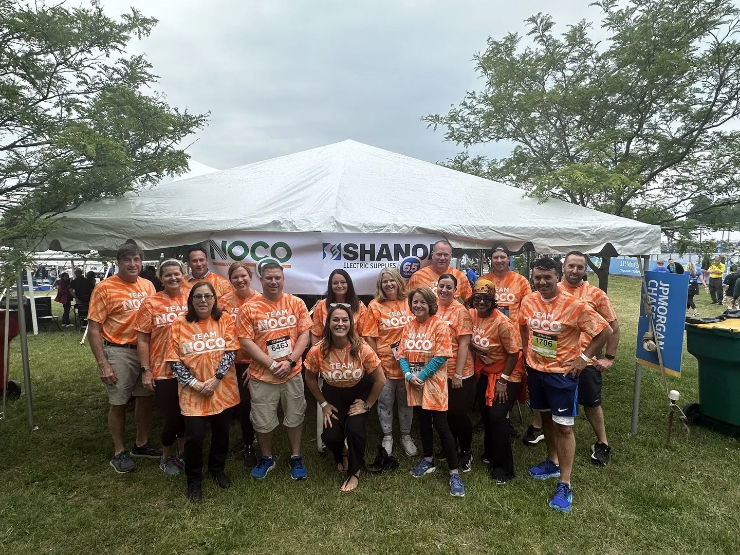 Group of people in orange Team NOCO shirts posing in front of a white event tent outdoors.