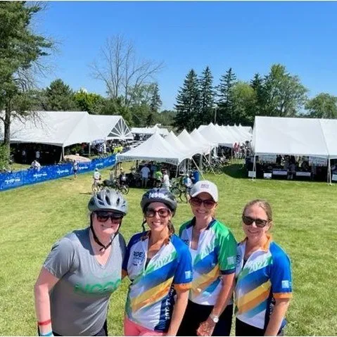 Four women in cycling gear smile in front of white event tents on a sunny grassy field.