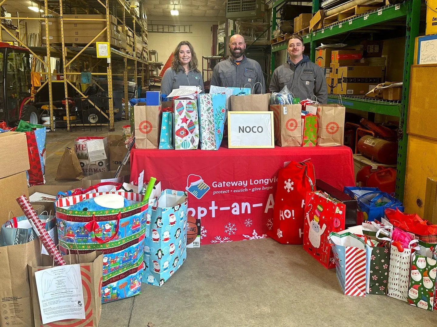 Three people stand behind a table with holiday gifts in a warehouse, smiling at the camera.