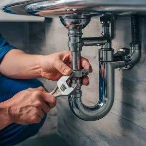 Person using a wrench to adjust plumbing pipes under a sink in a bathroom or kitchen.