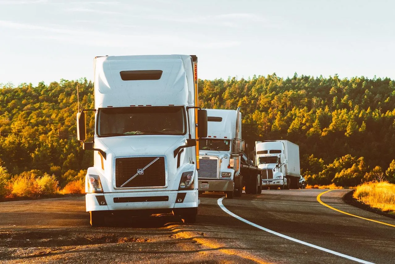 Three white semi-trucks parked on a curved road with trees and bushes in the background.