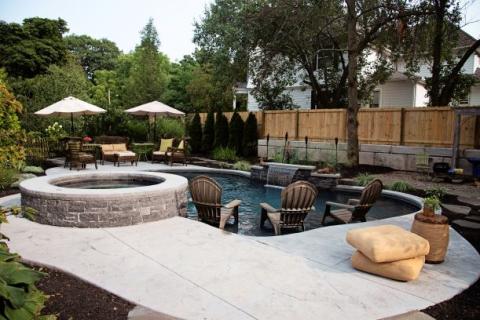 Backyard with a pool, hot tub, lounge chairs, tables with umbrellas, and lush green trees.
