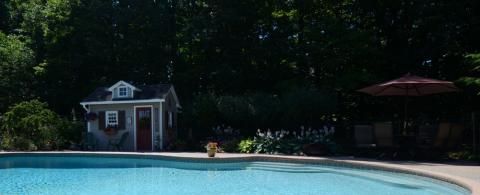 A backyard pool with a small shed and patio furniture under a red umbrella in the background.