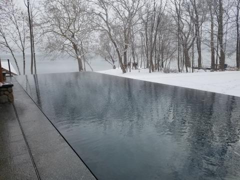 Infinity pool reflecting trees on a snowy, overcast day by a lake with bare trees in winter.