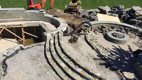 Stone patio and fire pit under construction with excavators and unfinished seating area.