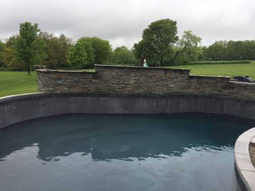 An empty backyard swimming pool with a stone wall, surrounded by grass and trees.