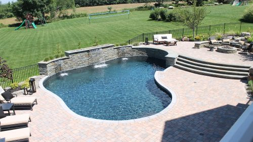 Curved backyard pool with stone waterfall, patio seating, and view of large grassy yard.