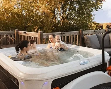 Four people, including two children, relax together in a hot tub on a deck surrounded by trees.