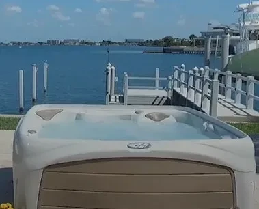 A hot tub sits near a dock overlooking a calm bay with boats and buildings in the distance.