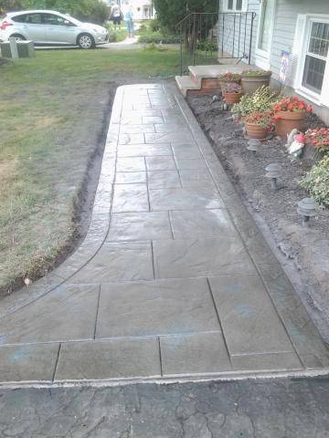 Freshly installed, stamped concrete walkway leading to a house entrance with flower pots nearby.