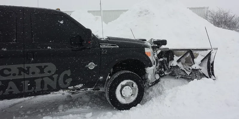 A black pickup truck with a snowplow clears deep snow in a snowy, winter setting.