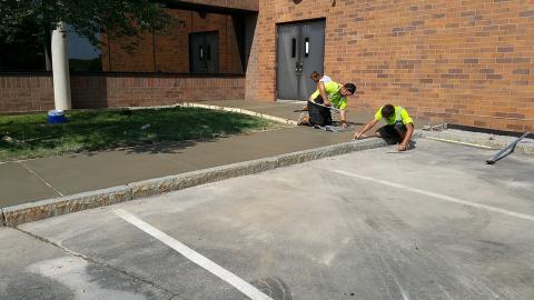 Two workers in yellow shirts smooth freshly poured concrete next to a brick building.
