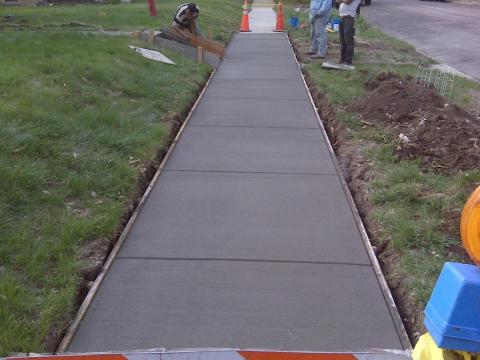 Workers smoothing wet concrete on a newly poured sidewalk bordered by grass and caution cones.
