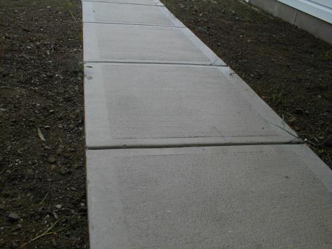 A concrete sidewalk with square sections runs beside bare soil and a building wall.