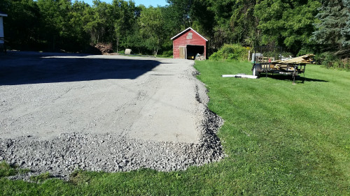 A gravel driveway leads to a small red barn, with grass and trees on both sides.