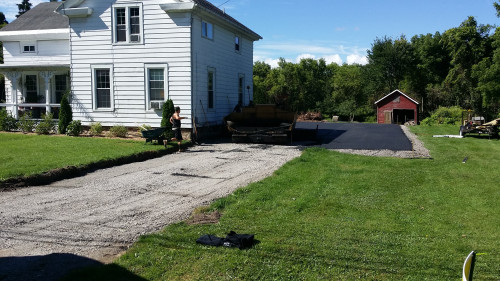 A crew paves a driveway with asphalt beside a white house and a red garage in a green yard.